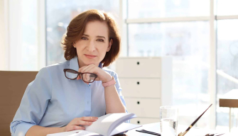 1. A woman wearing glasses sits at a desk, writing in a notebook, focused on her work.