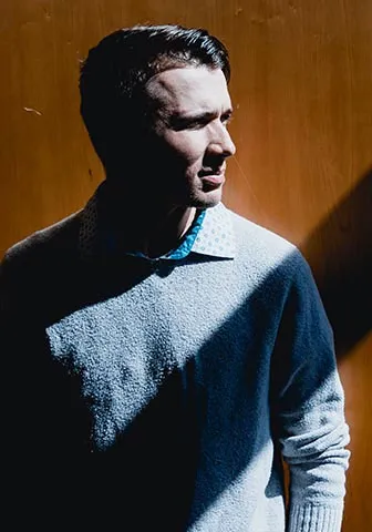 A man stands in front of a wooden door, looking directly at the camera with a neutral expression.