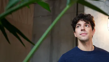 A man stands in front of a green plant, smiling and wearing a casual shirt, with a bright background.