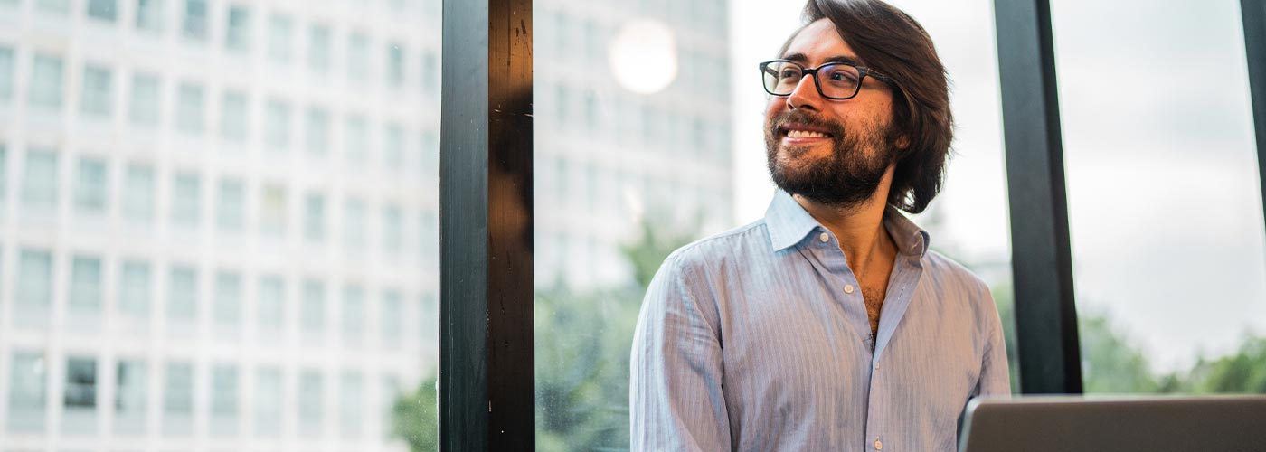 A man with glasses and a beard stands in front of a window, looking thoughtfully outside.