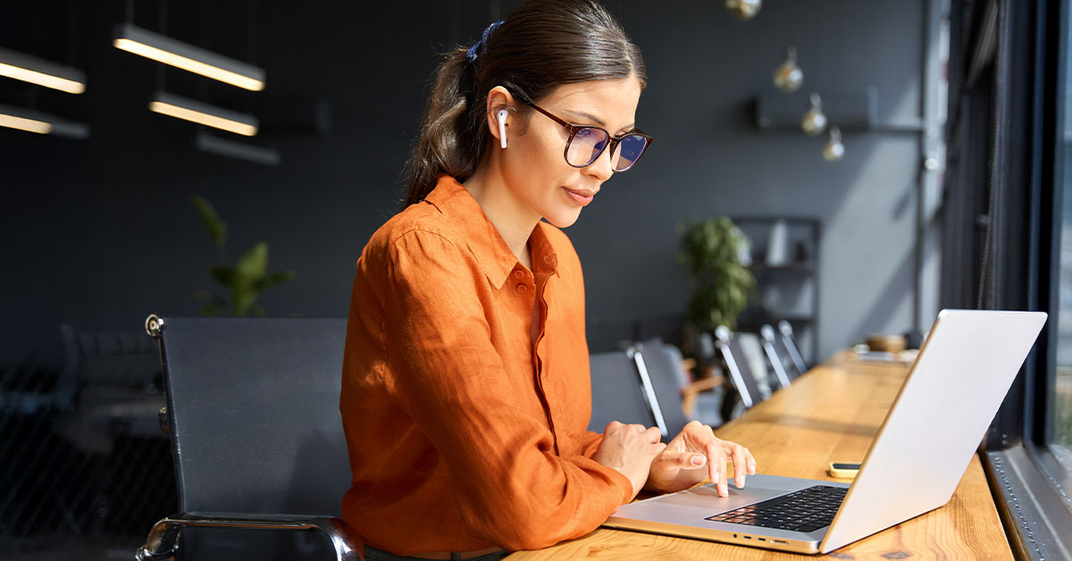 A woman wearing glasses is focused on her laptop, engaged in work at a desk.