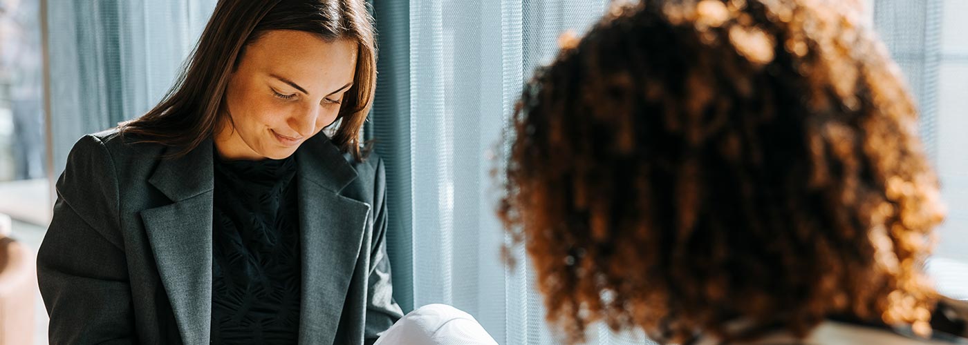 A woman speaks to another woman in an office setting, both engaged in a professional conversation.