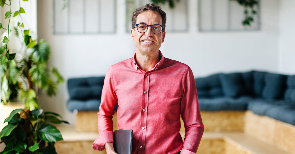 A man wearing glasses holds a book while standing in an office setting.